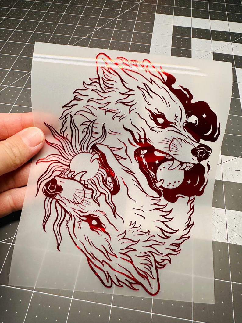 May include: A red wolf graphic with intricate details, featuring two wolf heads and a sun design. The artwork is on a transparent sheet, showcasing a detailed illustration suitable for various applications.