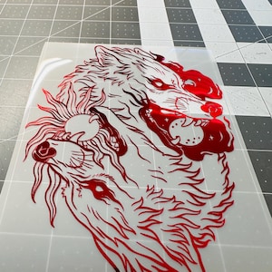 May include: Red vinyl decal featuring a detailed illustration of two wolves, one howling at a sun with a face. The design is a bold, graphic representation of the animals, with intricate line work and a metallic sheen.