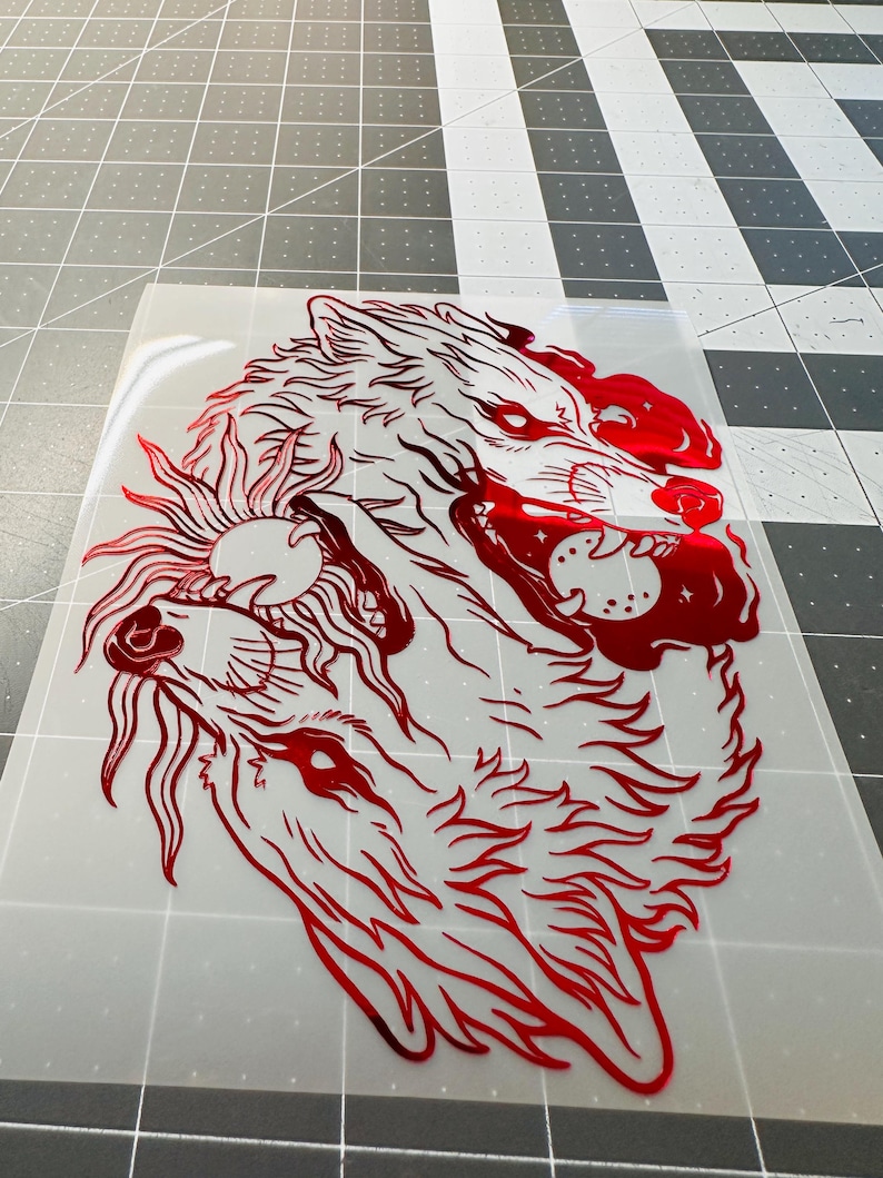 May include: Red vinyl decal featuring a detailed illustration of two wolves, one howling at a sun with a face. The design is a bold, graphic representation of the animals, with intricate line work and a metallic sheen.