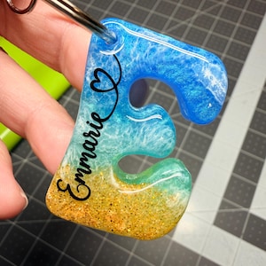 May include: A resin keychain in the shape of the letter "E", with a beach-themed design. The top is blue, transitioning to turquoise and gold glitter at the bottom. The name "Emmarie" is written in black script, with a heart symbol.
