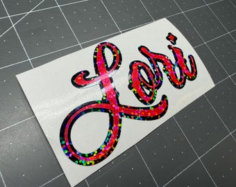 May include: A custom vinyl decal featuring the name Lori written in a bold, cursive script. The lettering has a vibrant red holographic finish with a multi-colored glitter effect, outlined in a thick black border on a white backing sheet.