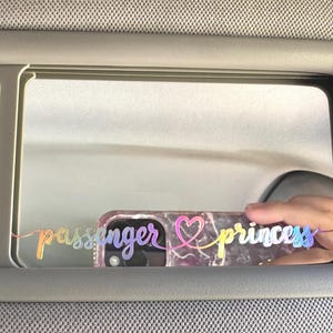May include: A car's rearview mirror with a rainbow iridescent sticker that reads "passenger princess" with a heart symbol.