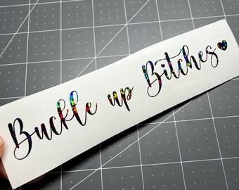 May include: A rectangular vinyl decal featuring the text "Buckle up Bitches" written in a black cursive script with a holographic, multi-colored glitter effect inside the letters, finished with a small black heart symbol at the end.