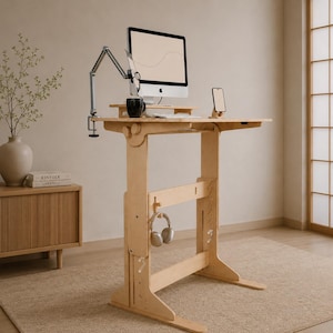 Pode incluir: Uma mesa de pé de madeira clara com um monitor, um braço ajustável prateado, uma caneca preta e um suporte para smartphone na superfície. Um par de auscultadores está pendurado num gancho na viga de suporte central da estrutura.