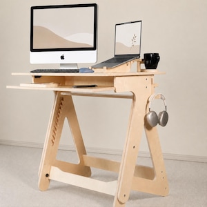 Pode incluir: Uma secretária de pé em madeira clara com design em forma de A, incluindo uma bandeja deslizante para teclado, um suporte para portátil e um gancho lateral para auscultadores. Contém um computador, um portátil, uma caneca preta e um rato.