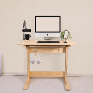 Peut inclure: Un bureau debout en bois clair doté d'une surface de travail, d'un cadre de support central et d'un crochet pour suspendre des accessoires. Le bureau supporte un écran d'ordinateur, un clavier noir, une tasse noire, un support de téléphone et un casque argenté.