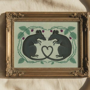Puede incluir: Una impresión artística enmarcada que presenta dos ratones negros enfrentados, con sus colas formando un corazón. La obra de arte está sobre un fondo verde claro con un borde de hojas verdes, espinas y bayas moradas. El marco es dorado.