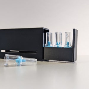 Könnte beinhalten: Ein schwarzes Plastikgehäuse mit einem transparenten Plastikfach, das fünf kleine, transparente, zylindrische Röhrchen mit blauen Spitzen enthält. Das Gehäuse ist geöffnet und eines der Röhrchen liegt auf einer weißen Oberfläche.