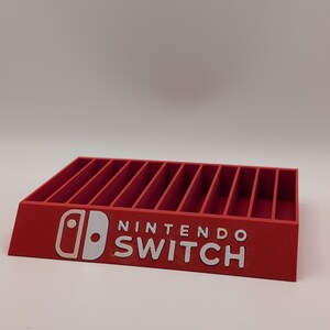 Könnte beinhalten: Ein roter Nintendo Switch-Spielkassettenhalter. Der Halter hat mehrere vertikale Schlitze, und das Nintendo Switch-Logo und der Text sind weiß. Der Halter besteht aus einem festen Material und ist für Spielkassetten konzipiert.