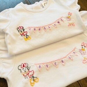 Puede incluir: Dos camisetas blancas para beb&eacute;s con personajes de dibujos animados bordados y nombres. Una camiseta presenta a Minnie Mouse y Daisy Duck con el nombre "CAROLINE" en una pancarta. La otra camiseta tiene a Minnie Mouse y Daisy Duck con el nombre "AVERY" en una pancarta.