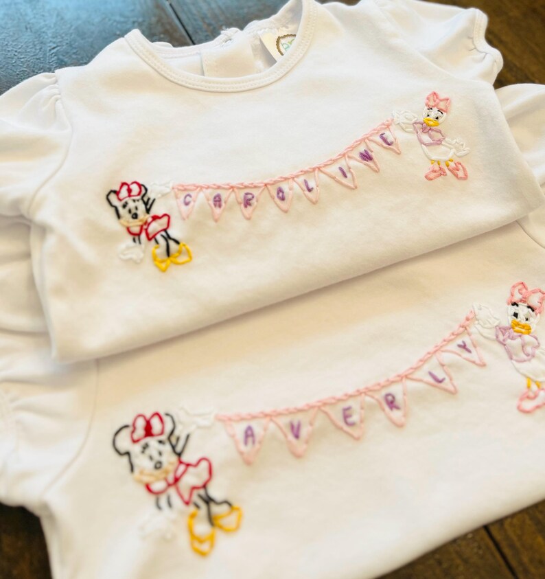 Puede incluir: Dos camisetas blancas para beb&eacute;s con personajes de dibujos animados bordados y nombres. Una camiseta presenta a Minnie Mouse y Daisy Duck con el nombre "CAROLINE" en una pancarta. La otra camiseta tiene a Minnie Mouse y Daisy Duck con el nombre "AVERY" en una pancarta.