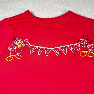 May include: Red t-shirt with embroidered Mickey Mouse and Donald Duck characters holding a banner that says "GEORGE".
