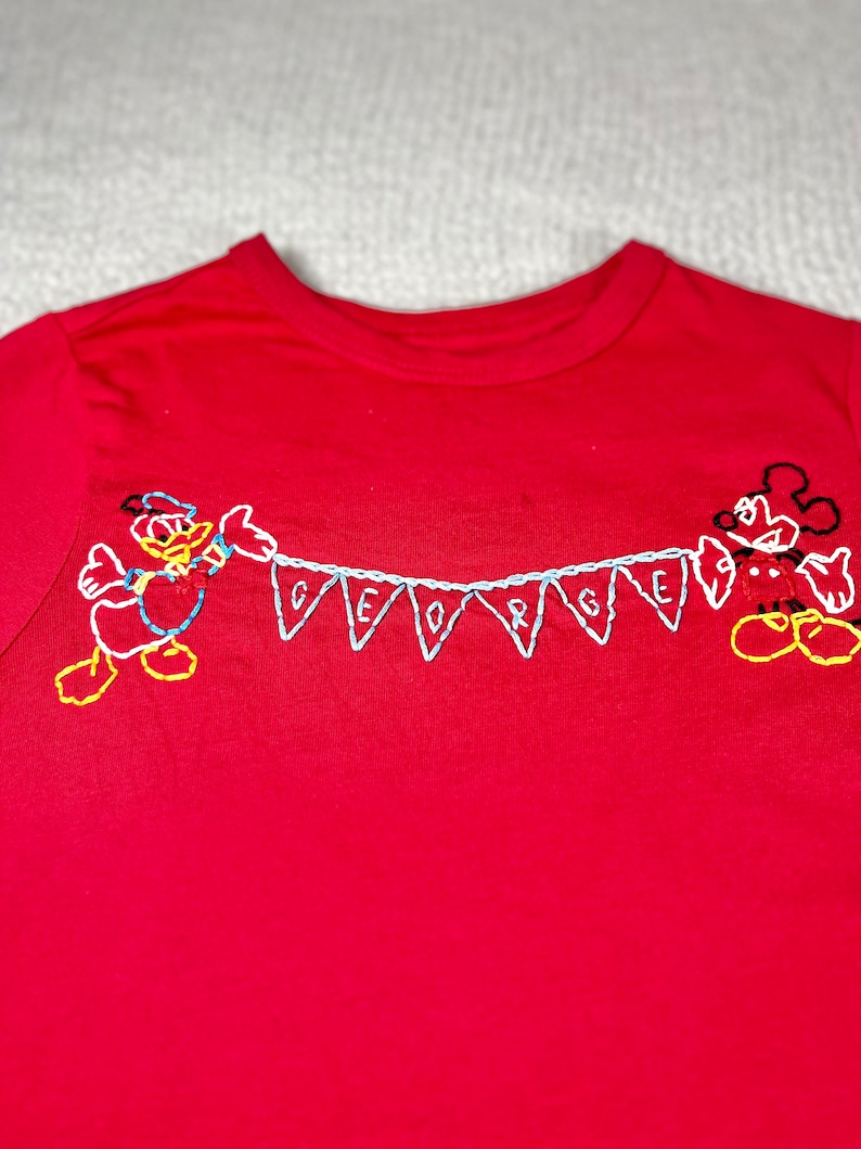 May include: Red t-shirt with embroidered Mickey Mouse and Donald Duck characters holding a banner that says "GEORGE".