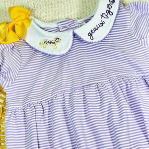 May include: A purple and white striped baby dress with a white Peter Pan collar. The collar has an embroidered tiger and the text "geaux tigers" in purple thread.