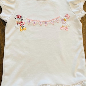 Puede incluir: Camiseta blanca de manga corta con volantes en las mangas y el dobladillo. Bordada con Minnie Mouse y Daisy Duck sosteniendo una pancarta rosa que dice "AVERLY". Un peque&ntilde;o logotipo circular en el cuello.