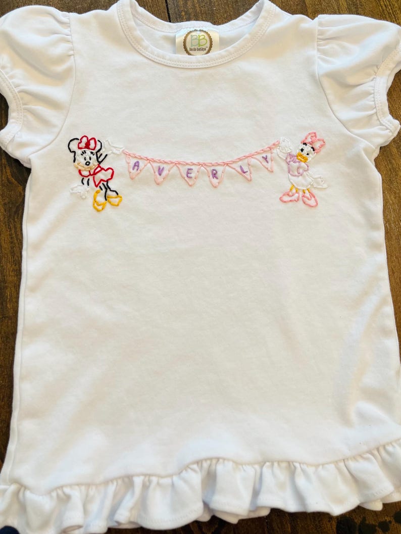 Puede incluir: Camiseta blanca de manga corta con volantes en las mangas y el dobladillo. Bordada con Minnie Mouse y Daisy Duck sosteniendo una pancarta rosa que dice "AVERLY". Un peque&ntilde;o logotipo circular en el cuello.