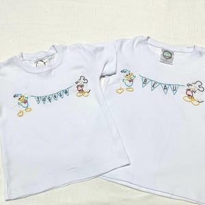 May include: Two white t-shirts with embroidered Mickey Mouse characters holding a banner that says "Tucker" and "Beau".