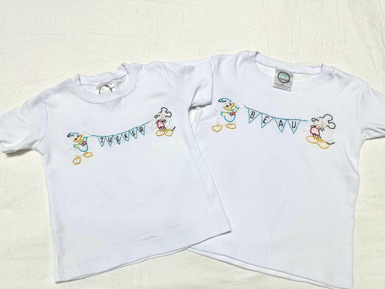 May include: Two white t-shirts with embroidered Mickey Mouse characters holding a banner that says "Tucker" and "Beau".
