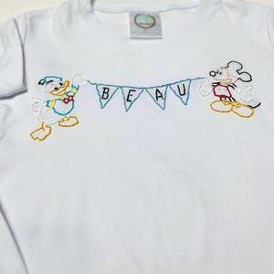 May include: White t-shirt with embroidered images of Mickey Mouse and Donald Duck holding a banner that reads "BEAU".
