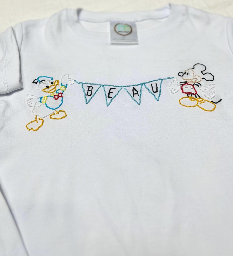 May include: White t-shirt with embroidered images of Mickey Mouse and Donald Duck holding a banner that reads "BEAU".