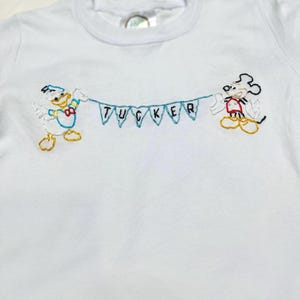 May include: White t-shirt with embroidered images of Mickey Mouse and Donald Duck holding a banner that says "TUCKER".