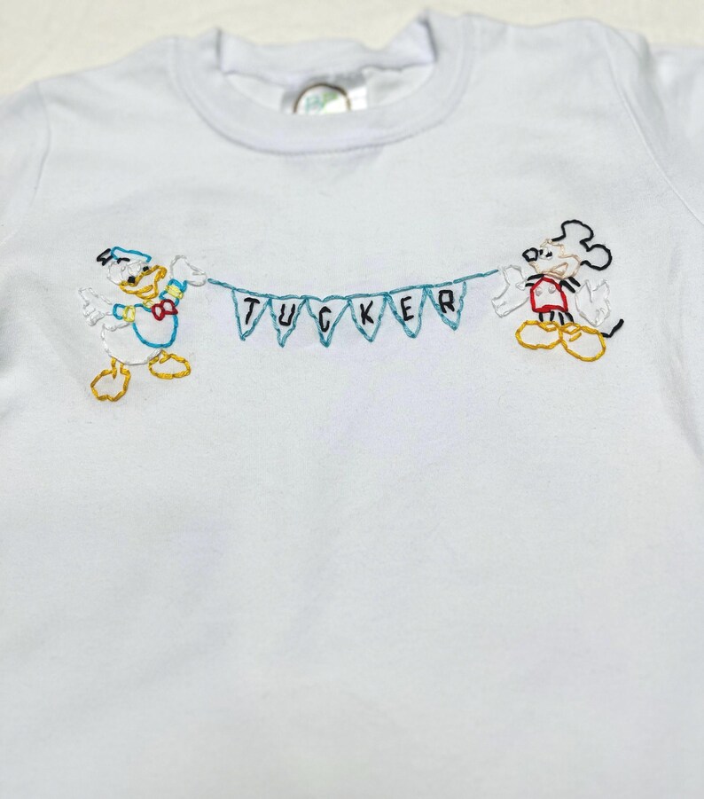 May include: White t-shirt with embroidered images of Mickey Mouse and Donald Duck holding a banner that says "TUCKER".