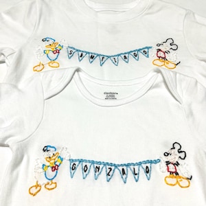 May include: Two white t-shirts with embroidered Disney characters. The top shirt has the name "SANTIAGO" embroidered on a blue banner held by Mickey Mouse and Donald Duck. The bottom shirt has the name "GONZALO" embroidered on a blue banner held by Mickey Mouse and Donald Duck.