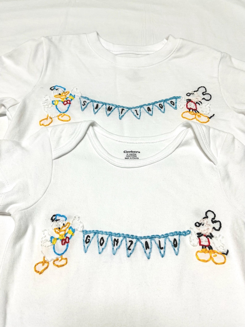 May include: Two white t-shirts with embroidered Disney characters. The top shirt has the name "SANTIAGO" embroidered on a blue banner held by Mickey Mouse and Donald Duck. The bottom shirt has the name "GONZALO" embroidered on a blue banner held by Mickey Mouse and Donald Duck.