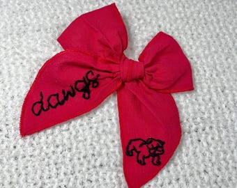 May include: A bright pink fabric bow with the word "dawgs" embroidered on one side and a black embroidered bulldog on the other side.