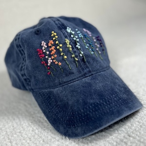 Può includere: Un berretto da baseball in denim blu scuro con un motivo di fiori ricamati arcobaleno.