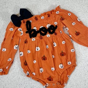 May include: Orange baby onesie with a white pumpkin pattern and a black felt "boo" applique. The onesie has long sleeves and a ruffled neckline. A black bow is included.