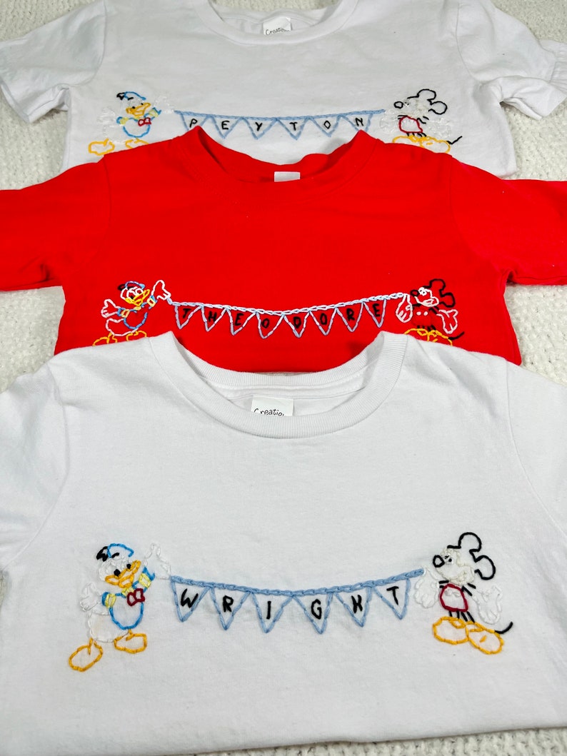 May include: Three white t-shirts with embroidered Disney characters and names. The top shirt has Mickey Mouse and the name Peyton. The middle shirt has Donald Duck and the name Theodore. The bottom shirt has Donald Duck and Mickey Mouse and the name Wright.