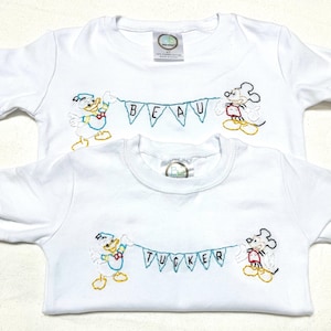 May include: Two white t-shirts with embroidered images of Mickey Mouse and Donald Duck holding a banner. The top shirt has the name "BEAU" on the banner. The bottom shirt has the name "TUCKER" on the banner.