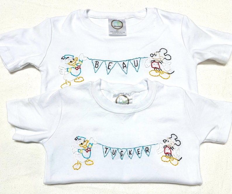 May include: Two white t-shirts with embroidered images of Mickey Mouse and Donald Duck holding a banner. The top shirt has the name "BEAU" on the banner. The bottom shirt has the name "TUCKER" on the banner.