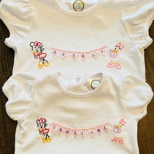 Puede incluir: Camisetas blancas de manga corta con mangas con volantes. Cada camiseta presenta personajes de dibujos animados bordados, una pancarta con un nombre y un logotipo. La camiseta superior dice "AVERY" y la inferior dice "CAROLINE".