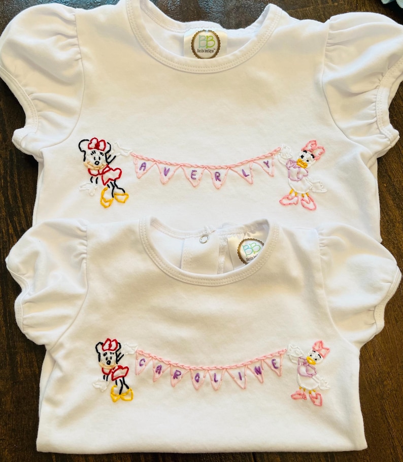 Puede incluir: Camisetas blancas de manga corta con mangas con volantes. Cada camiseta presenta personajes de dibujos animados bordados, una pancarta con un nombre y un logotipo. La camiseta superior dice "AVERY" y la inferior dice "CAROLINE".