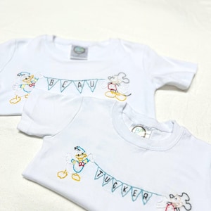 May include: Two white t-shirts with embroidered Disney characters, Donald Duck and Mickey Mouse, holding a banner with the names "Beau" and "Tucker" on them.