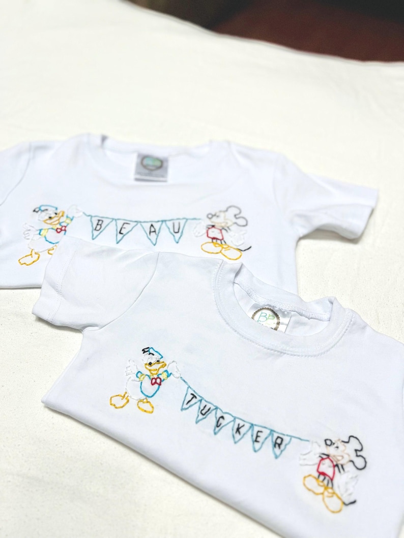 May include: Two white t-shirts with embroidered Disney characters, Donald Duck and Mickey Mouse, holding a banner with the names "Beau" and "Tucker" on them.
