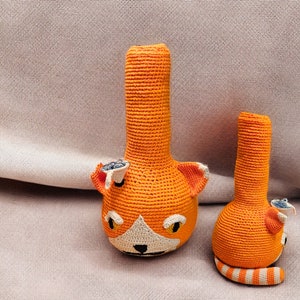 Puede incluir: Dos pipas de agua de ganchillo de color naranja y blanco con forma de gato. La pipa más grande tiene una cara de gato y la más pequeña tiene una cola rayada.