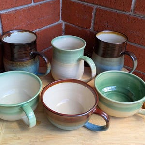 May include: A selection of handmade ceramic mugs and bowls in various sizes and colours. The mugs feature brown, blue, and green glazes, while the bowls have matching colour schemes. The pottery is arranged on a wooden surface against a brick wall.