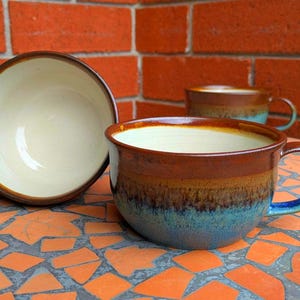 May include: Three ceramic soup bowls in brown, blue, and cream tones. The bowls have handles and are set on a mosaic tile surface. A red brick wall forms the backdrop.