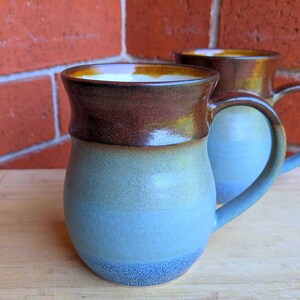 May include: Two ceramic mugs with a blue and brown glaze. The mugs have a rounded shape with a handle and are filled with a white liquid. The background features a red brick wall and a wooden surface.