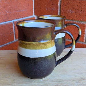 May include: Two ceramic mugs with a brown, gold, and white striped design. The mugs have a dark brown base and interior, with a white stripe and a gold stripe. The handles are black with a white and gold accent.