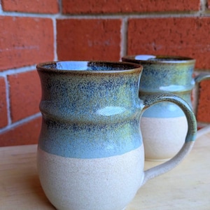 May include: Two ceramic mugs in blue and beige. The mugs have a rounded shape with a handle and a textured surface. The top of the mugs are a dark blue, transitioning to a lighter blue, and then beige at the bottom.