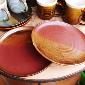 May include: Two ceramic plates with a reddish-brown and tan gradient. The plates are round and have a slightly raised edge. Several mugs and saucers in shades of blue, brown, and tan are also visible, suggesting a tableware set.