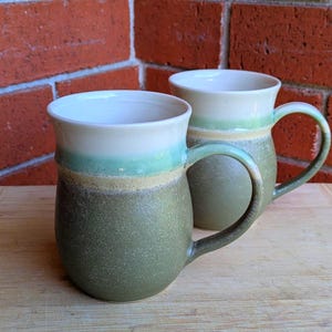 May include: Two handmade ceramic mugs with a textured, speckled finish. The mugs feature a gradient of colours, including green, tan, and white. Each mug has a rounded body and a curved handle, perfect for enjoying a warm drink.