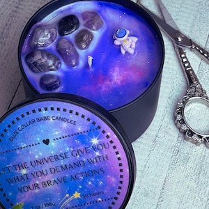 May include: A galaxy-themed candle with a black lid and a purple and blue wax design. The candle has a small astronaut figurine and amethyst crystals embedded in the wax. The lid has a quote that reads "Let the universe give you what you demand with your brave actions." The lid also has a star design and the text "7 oz/198g, 30 hours, Be The Change."