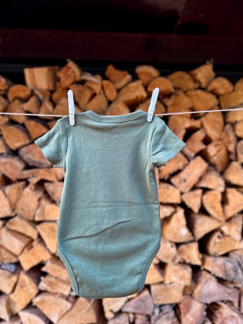 Puede incluir: Un pelele de beb&eacute; verde colgado de una cuerda para tender la ropa frente a una pila de le&ntilde;a apilada.