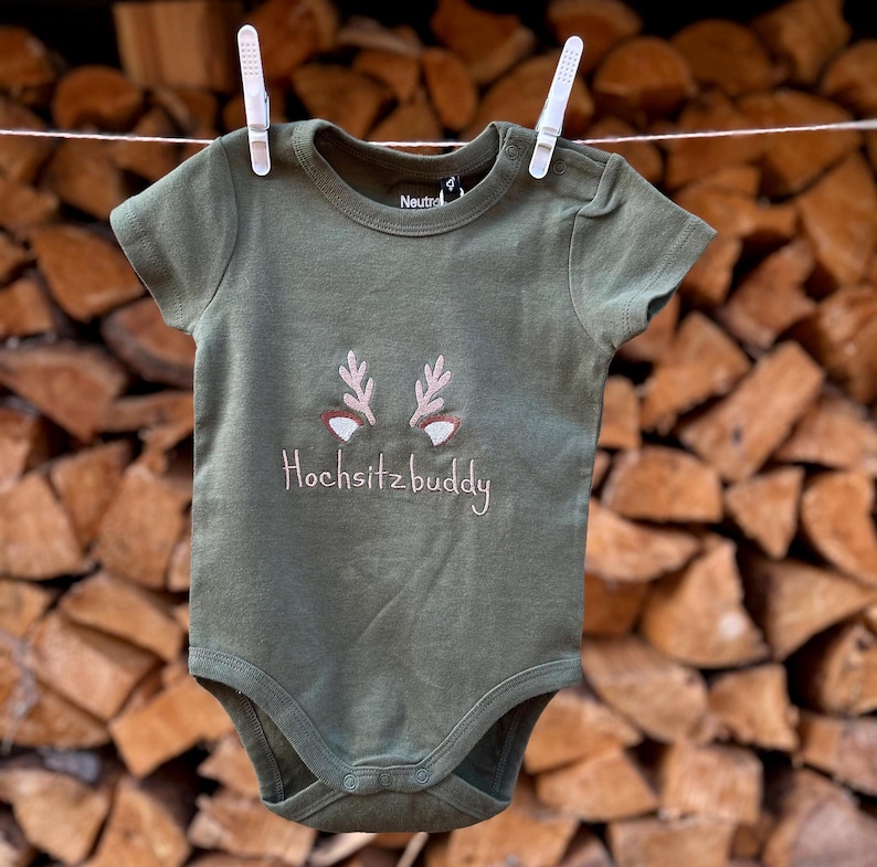 Puede incluir: Un body de beb&eacute; verde con una cara de ciervo bordada en la parte delantera. El body dice "Hochsitzbuddy" en bordado rosa.