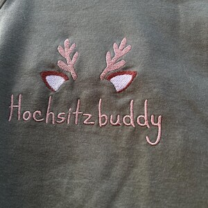 Puede incluir: Una camiseta verde de manga larga con cuernos de reno bordados en rosa y el texto "Hochsitzbuddy".
